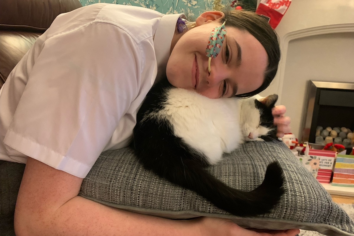 A teenage girl with a tube going into her nose cuddles a black and white cat