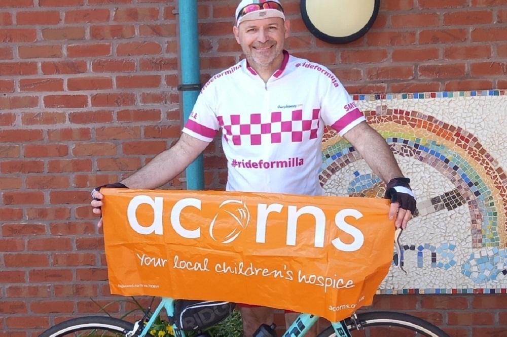 A man wearing a cycling jersey with the slogan ride for milla. He is standing behind a bicycle and holding an orange Acorns banner